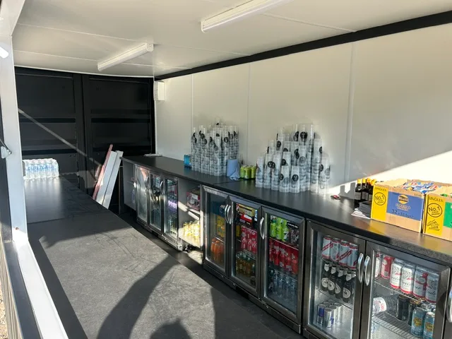 Inside a container bar — dark bar counter with a row of glass-front under-counter bottle fridges stocked with drinks