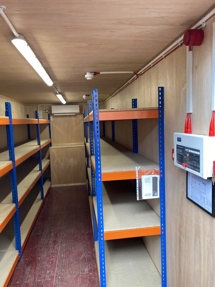 Lined interior of a 20ft battery storage container with racking, LED strip lighting, air conditioning and a fire-alarm panel